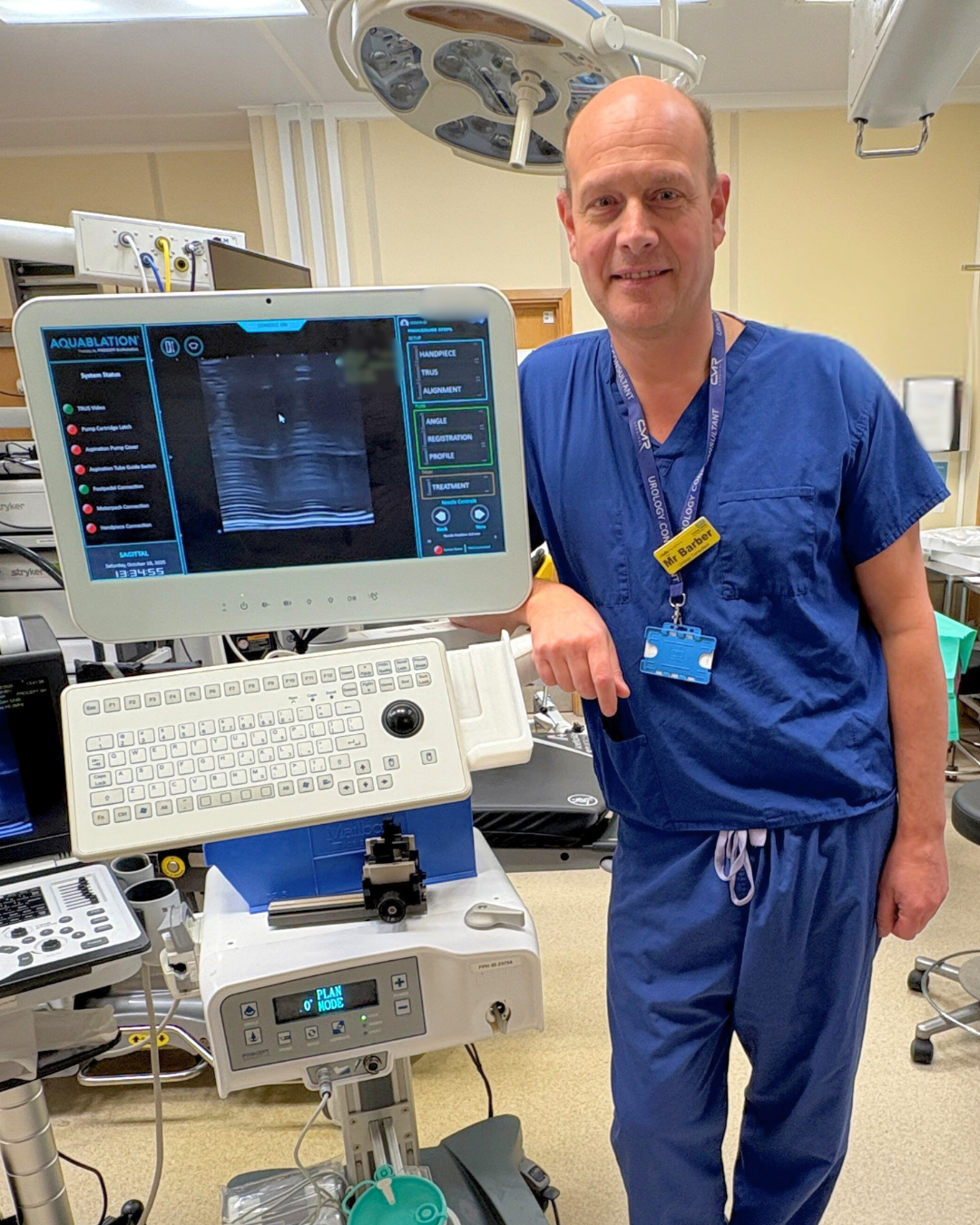 Photo of Neil Barber wearing scrubs in theatre, leaning on a stand with medical devices