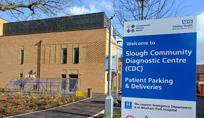 A footpath leading to a new building in yellow brick, showing cycle parking. A sign is visible directing people to the Slough CDC, including a message that the nearest emergency department is at Wexham Park Hospital.