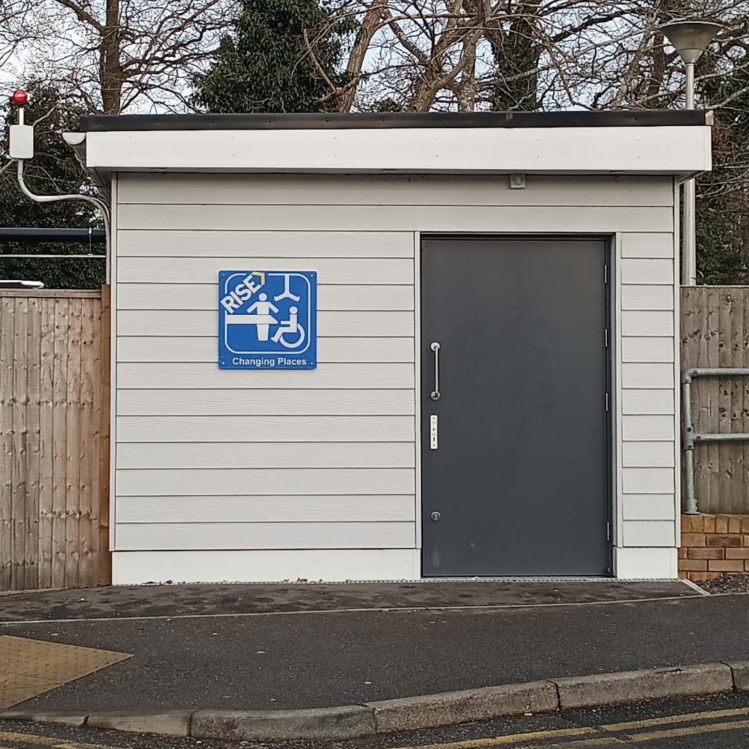 Photo of changing places toilet. A small outbuilding set back from the road with a paved footpath in between. Sign with changing places branding.